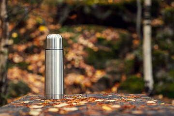 Steel thermos with delicious hot tea in the autumn forest on an old wooden table with fallen leaves...
