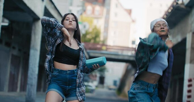 Hip-hop street dance by two teenage girls dressed in comfortable clothes. The girls hold a hand-held speaker from which music flies to their choreography.