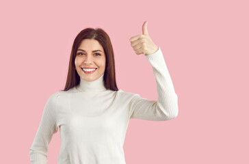 Beautiful positive Caucasian woman showing thumbs up and smiling broadly agreeing to promotional offer demonstrating approval dressed in white casual turtleneck stands on plain pink background