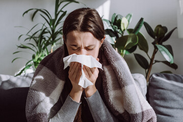 Sick frozen young female seated on sofa in living room covered with warm cozy plaid sneezing holding paper napkin blow out runny nose feels unhealthy, seasonal cold. Weakened immune system concept. - obrazy, fototapety, plakaty