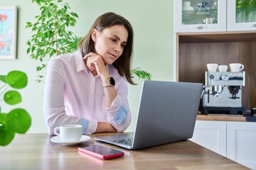 Young woman sitting at home using laptop, drinking morning coffee