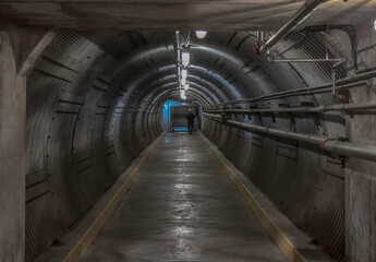Long underground mountain tunnel with entrance in sight, corrugated metal walls, concrete floors, piping, nobody