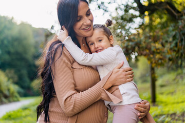 Fototapeta premium Happy pregnant mother and daughter hugging on a park bench. Happy motherhood. Beautiful woman with a little daughter in nature