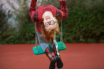 Outdoor porrait of funny little girl playing on swing, looking upside down, wearing glasses