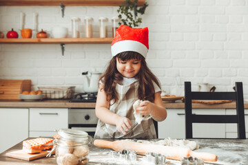 Portrait of a happy little girl in a Santa hat, rolling out dough on the kitchen table, a child preparing Christmas cookies.