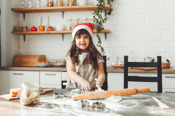 Portrait of a happy little girl in a Santa hat, rolling out dough on the kitchen table, a child preparing Christmas cookies.