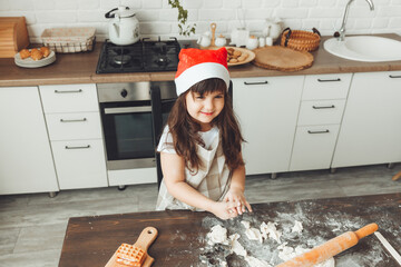 Portrait of a happy little girl in a Santa hat, rolling out dough on the kitchen table, a child preparing Christmas cookies.