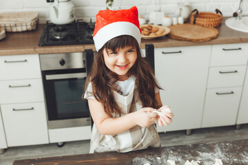Portrait of a happy little girl in a Santa hat, rolling out dough on the kitchen table, a child preparing Christmas cookies.