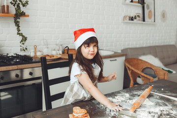 Portrait of a happy little girl in a Santa hat, rolling out dough on the kitchen table, a child preparing Christmas cookies.