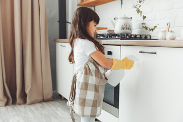 A cute little girl in an apron wipes the cupboard in the kitchen, washes with a sponge in a...