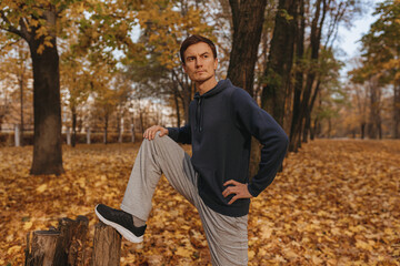 Fototapeta premium Confident sporty male athlete stretching legs leaning on tree while having fitness training outdoors in autumn park 