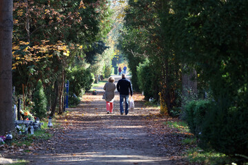 Starsi ludzie spacerują jesienną alejką na cmentarzu. 