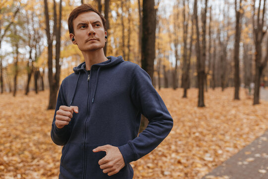 Confident male athlete in activewear running in autumn park and listening to music in wireless earbuds during outdoor training while leading healthy lifestyle  