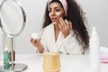Cheerful tanned curly Latin lady apply facial cream at cheeks admiring herself in mirror doing spa procedure in home interior. Copy space. Aesthetic medicine skincare beauty products ad concept