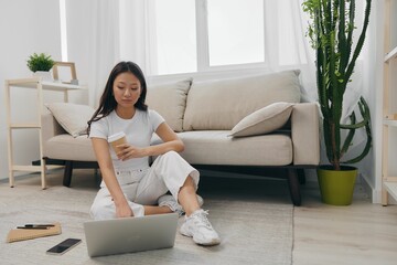 Young Asian female student looking at laptop screen sitting on floor at home with notebooks to write, lifestyle work freelance