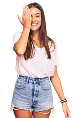 Young hispanic woman wearing casual white tshirt covering one eye with hand, confident smile on face and surprise emotion.