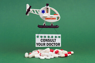 On a green surface, an ambulance helicopter, pills and a white sign with the inscription - Consult your doctor