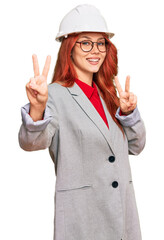 Young redhead woman wearing architect hardhat smiling looking to the camera showing fingers doing victory sign. number two.