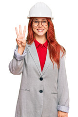 Young redhead woman wearing architect hardhat showing and pointing up with fingers number three while smiling confident and happy.