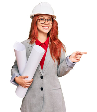 Young redhead architect woman wearing safety helmet holding blueprints smiling happy pointing with hand and finger to the side