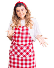 Young caucasian woman wearing apron clueless and confused expression with arms and hands raised....