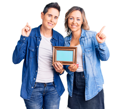 Couple of women holding empty frame with a big smile on face, pointing with hand finger to the side looking at the camera.
