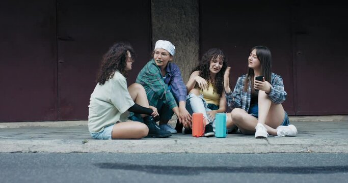 A crew of girl friends sits on the sidewalk in the city dressed in casual streetwear clothes. The crazed teens take pictures of themselves, record a video for social media.
