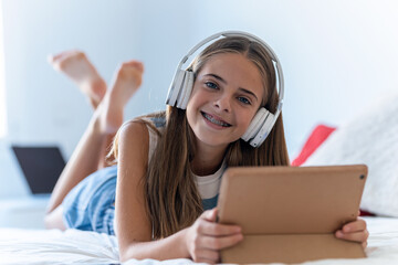 Beautiful young girl listening to music with headphones while playing with digital tablet lying on the bed.
