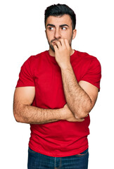 Hispanic man with beard wearing casual red t shirt looking stressed and nervous with hands on mouth biting nails. anxiety problem.