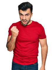 Hispanic man with beard wearing casual red t shirt angry and mad raising fist frustrated and...