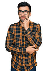 Hispanic man with beard wearing casual clothes and glasses serious face thinking about question with hand on chin, thoughtful about confusing idea