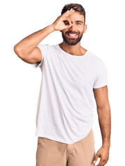 Young hispanic man wearing casual white tshirt doing ok gesture with hand smiling, eye looking through fingers with happy face.