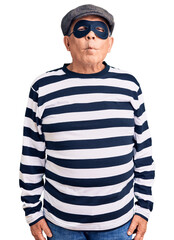 Senior handsome man wearing burglar mask and t-shirt making fish face with lips, crazy and comical...