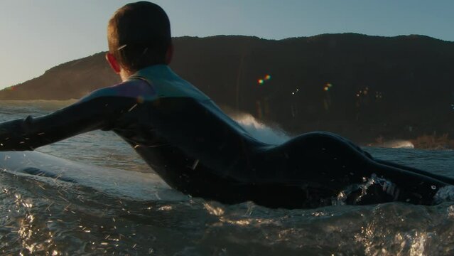 Boy surfer paddles in the ocean at sunrise and watches another surfer riding the wave