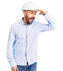 Young handsome man wearing architect hardhat very happy and smiling looking far away with hand over head. searching concept.