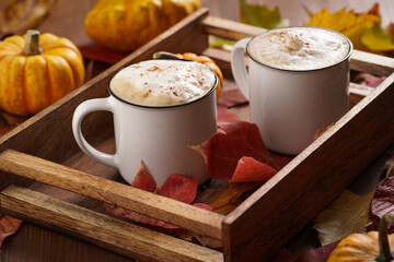 Two white cups with black coffee and white milk foam and cinnamon spice in a cozy autumn setting with red, yellow and orange leaves, hokkaido pumpkins, on wooden background