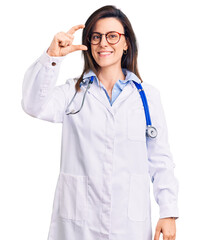 Young beautiful woman wearing doctor stethoscope and glasses smiling and confident gesturing with hand doing small size sign with fingers looking and the camera. measure concept.