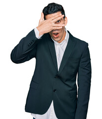 Handsome hispanic man wearing business clothes peeking in shock covering face and eyes with hand, looking through fingers with embarrassed expression.