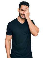 Hispanic man with beard wearing casual black t shirt covering one eye with hand, confident smile on face and surprise emotion.