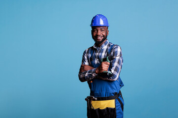 Optimistic african american builder using tool belt and cordless power drill. Contractor proud of skills looking confidently at camera in studio isolated on blue background.