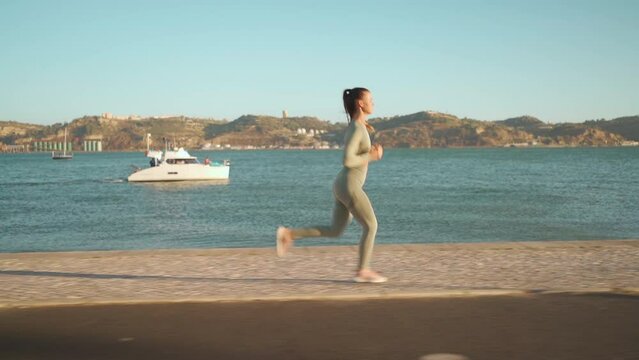 Confident fit female jogging by embankment area coastal city in beautiful sunny day do fitness training for cardio health. Tracking shot. Slow motion. Handheld. Athletic woman running, morning run