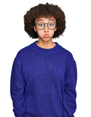 Young hispanic girl wearing casual winter sweater and glasses puffing cheeks with funny face. mouth inflated with air, crazy expression.