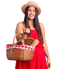 Young beautiful brunette woman wearing summer hat holding picnic basket looking positive and happy...