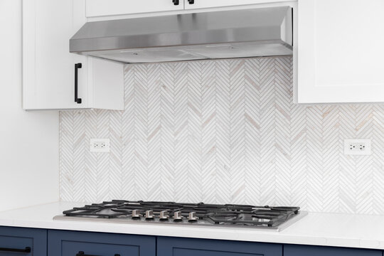 A detail shot of a white and blue kitchen's stainless steel stove top, granite countertop, and a marble herringbone tiled backsplash.