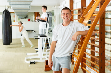 Portrait of cheerful sporty adult man posing during workout in gym, standing leaning against wall bars