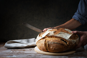 Traditional loaf of bread in the hands of a baker with cutting board and knife on floured pastry board, space for text.