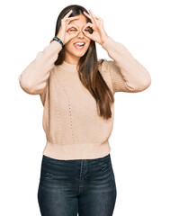 Young brunette woman wearing casual winter sweater doing ok gesture like binoculars sticking tongue out, eyes looking through fingers. crazy expression.