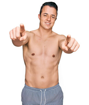 Handsome young man wearing swimwear shirtless pointing to you and the camera with fingers, smiling positive and cheerful