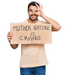 Young hispanic man holding mother nature is crying protest cardboard banner smiling happy doing ok sign with hand on eye looking through fingers