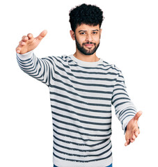 Naklejka premium Young arab man with beard wearing casual striped sweater looking at the camera smiling with open arms for hug. cheerful expression embracing happiness.